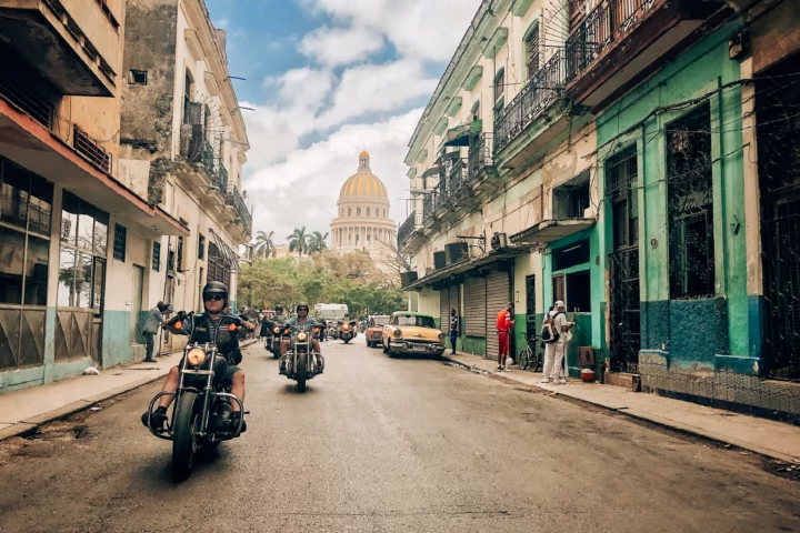 Motorradfahrer auf Harley-Davidsons durchqueren eine belebte Straße in Havanna, vorbei an klassischen Gebäuden mit der Kuppel des Kapitols im Hintergrund.